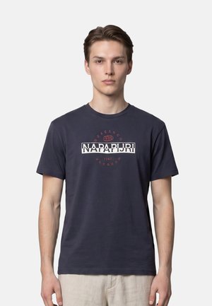 Jeune homme portant un t-shirt bleu foncé Napapijri avec texte logo, debout devant un fond uni gris clair.
