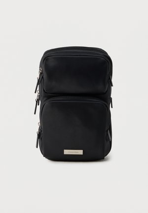 Bolso bandolera rectangular negro con dos compartimentos con cremallera y una pequeña placa metálica con el logo de Calvin Klein en el bolsillo frontal.