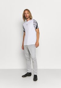 Witte sport t-shirt met grijs camouflage-patroon op de mouwen, gecombineerd met lichtgrijze trainingsbroeken en zwarte sportschoenen. Zwart logo-detail op de borst.