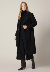 Długi czarny wełniany żakiet o podwójnym zapięciu, ze złotymi guzikami i paskiem w talii. Noszony na czarnym golfie i szerokich czarnych spodniach.