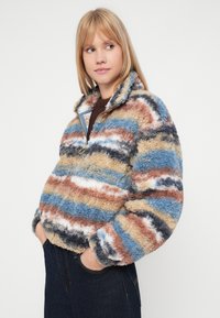 Suéter de polar difuso y multicolor con media cremallera, que presenta rayas horizontales en azul, marrón, beige y blanco, con una textura suave.