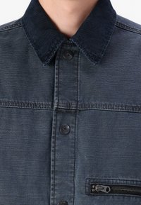 Dark denim jacket featuring a ribbed collar, button closure, and a zippered chest pocket, with a textured finish and contrasting stitching.
