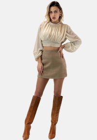 Blusa beige a maniche lunghe con tessuto arricciato; gonna beige chiara; stivali marroni al ginocchio con punta affilata; texture lisce in tutto.