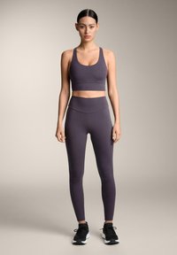 Ljubičasti sportski grudnjak i hlače visoki struk od rastezljivog materijala. Imaju glatku teksturu i minimalistički dizajn. Crne atletske cipele.