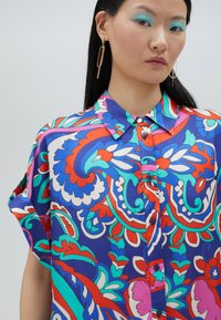 Blusa colorida con un patrón floral en azul, rojo y verde, mangas cortas, cierre de botones en la parte delantera y textura de tela suave.