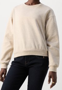 Sudadera beige con cuello redondo, puños y dobladillo acanalados. Textura suave, ajuste relajado, combinada con vaqueros oscuros. Patrón moteado sutil.