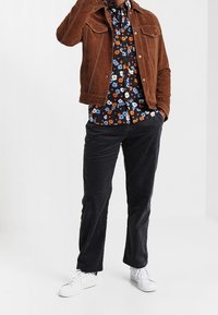 Chaqueta de ante marrón con costuras blancas, camisa de flores en azul y naranja, pantalones de pana negros y zapatillas blancas.