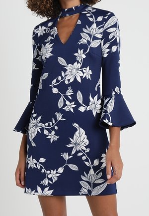 Vestito blu navy con motivo floreale bianco, scollo a goccia con fiocco al colletto e maniche svasate a tre quarti indossato da una persona.