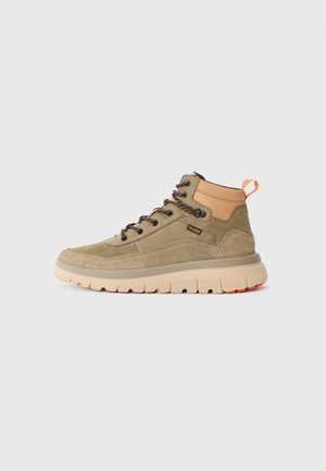 Light brown mid-top sneaker made of suede with a beige sole. Features black laces, a brown leather heel, and an orange pull tab.