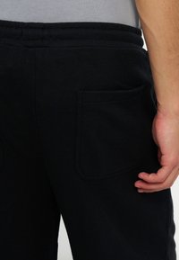 Gros plan d’une personne portant un pantalon de jogging noir avec une poche arrière et une chemise grise rentrée, montrant sa main droite à côté de la poche.