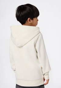 Niño con cabello corto y oscuro, vestido con una sudadera color crema, de perfil mirando hacia la derecha contra un fondo sencillo.