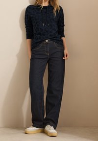 Felpa a stampa leopardo navy con cordino, abbinata a jeans a gamba larga blu scuro e sneakers bianche con suola gialla.