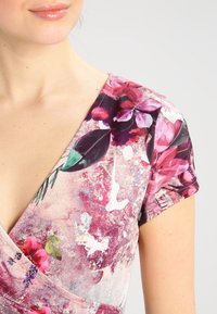 Närbild av en kvinna i en rosa blommig omlottklänning med korta ärmar och blandade botaniska mönster på texturerad tyg.