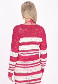 Långärmad, öppet stickad tröja med rosa och vita horisontella ränder. Har en ribbad nederkant och en detalj med knytning på baksidan. Lätt och texturerad.
