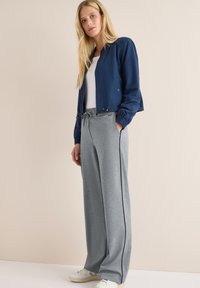 Femme blonde portant une veste marine, un haut blanc, un pantalon de survêtement gris à large jambe avec une bande noire, et des baskets blanches, se tenant devant un fond beige.
