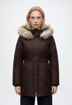 Femme portant un manteau d'hiver marron foncé avec une capuche bordée de fourrure, debout devant un fond blanc uni.