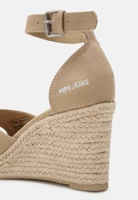 Sandale compensée beige avec une sangle en tissu au niveau de la cheville et un talon en jute tressé. Présente "PEPE JEANS" brodé en blanc à l'arrière. Texture lisse.