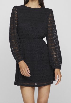 Kobieta w czarnej, koronkowej mini sukience z długim rękawem i zwężeniem w talii, stojąca na jasnoszarym tle.