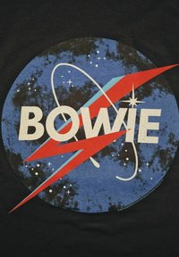 Grafický motiv s nápisem „BOWIE“ ve velkých bílých písmenech na kruhovém modrém pozadí, s červenými prvky ve tvaru blesku a motivy hvězd.