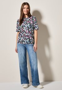 Korte mouwen, multikleurige top met abstract patroon, gecombineerd met wijde spijkerbroek in licht denim en witte sneakers op een neutrale achtergrond.