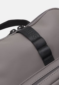 Grauer wasserdichter Beutel mit glatter Oberfläche, schwarzen Riemen-Details und kontrastierenden Nähten entlang der Reißverschlusslinie. Robuste Griffe.