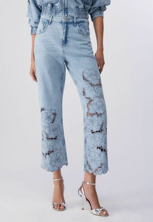 Svetlo modre kratke kavbojke z velikimi cvetličnimi čipkastimi izrezi in valovitimi robovi, nošene s srebrnimi sandalami z visokimi petami.