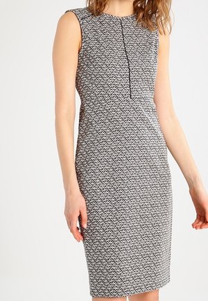 Ærmeløs knælængde kjole i sort og hvidt mønster med høj hals og diskrete folder foran, båret af en stående kvinde.