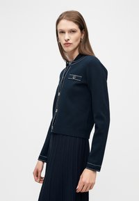 Γυναίκα που φοράει ένα κοντό σακάκι σε ναυτικό μπλε με λευκά ράμματα και κουμπιά, συνδυασμένο με μια αντίστοιχη πλισέ φούστα, στέκεται μπροστά σε ένα απλό φόντο.