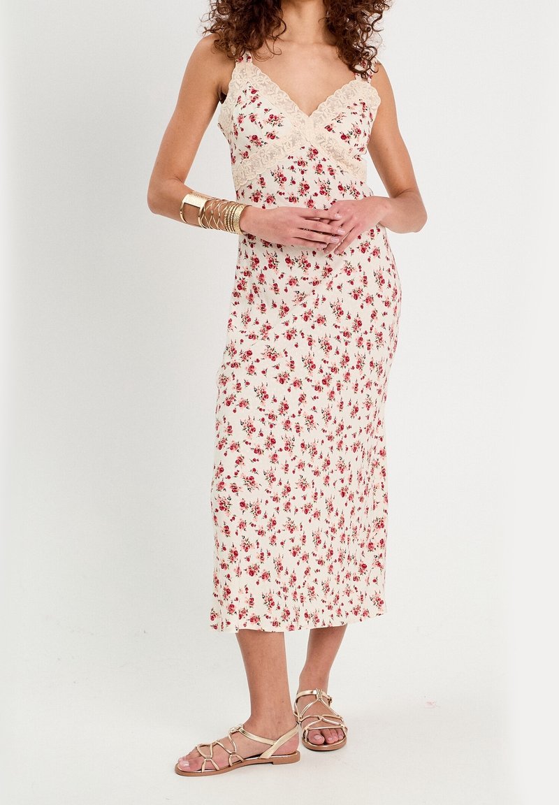 Femme portant une robe crème sans manches avec un petit imprimé floral rouge et une bordure en dentelle, des bracelets dorés et des sandales plates à brides.