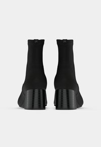 Pár černých kotníkových bot na podpatku, zobrazených ze zadní strany na jednobarevném bílém pozadí.