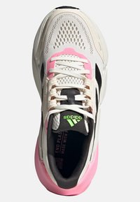Chaussure de sport blanche avec une tige en maille, accents rose, bande latérale noire et logo vert. Elle dispose de lacets et d'une semelle rembourrée.