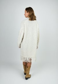 Frau mit dem Rücken zur Kamera, die einen knielangen, cremefarbenen Strickcardigan mit Fransen und hellbraunen Wildleder-Cowboystiefeln trägt, vor einem schlichten, hellen Hintergrund.