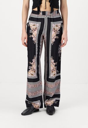 Femme portant un pantalon noir à jambes larges avec des motifs floraux et géométriques roses et beiges, associé à des chaussures noires à bout carré.