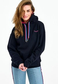 Mladá žena s volně rozpuštěnými blond vlasy, která nosí černou mikinu s růžovými a modrými šňůrkami a světle modré džíny s růžovými pruhy po stranách.