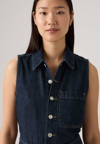 Ermeløs denimkjole i mørkeblått med klassisk krage, frontknapper og lomme på siden. Stoffet har en glatt tekstur med sømdetaljer.