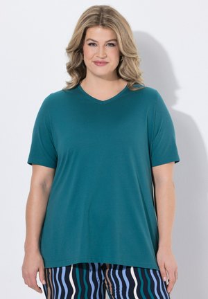 Olkapäille ulottuvat vaaleat hiukset omaava nainen, joka pukeutuneena sinivihreään lyhythihaiseen yläosaan ja kuvioituihin aaltoileviin raidallisiin housuihin, seisoo valkoista taustaa vasten.