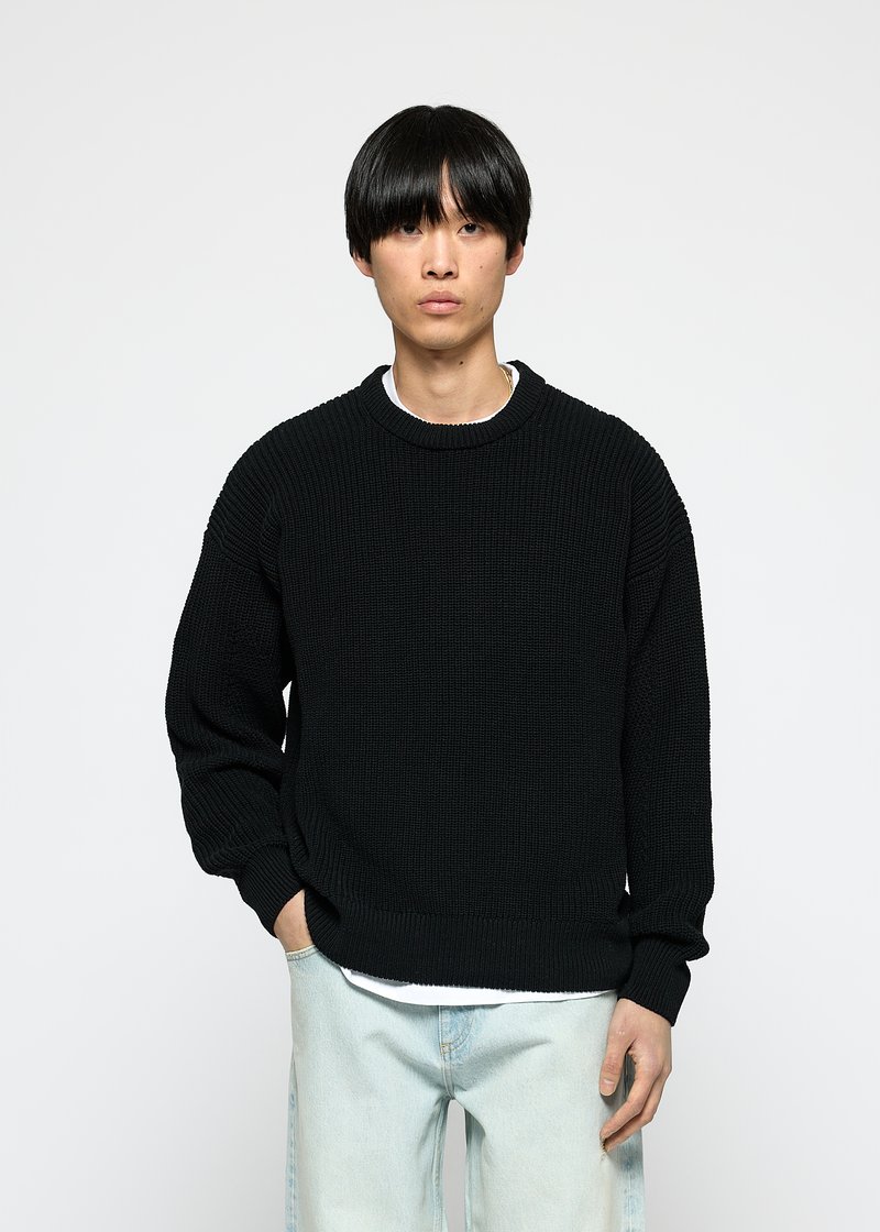 Junger Mann mit glattem schwarzen Haar, der einen schwarzen Strickpullover über einem weißen Hemd und hellblauen Jeans trägt, steht mit einer Hand in der Tasche.