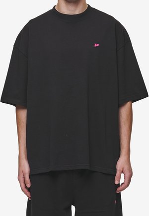Młoda osoba nosząca oversize czarną koszulkę z małym różowym logo litery P na górnej lewej części klatki piersiowej.