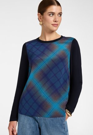 Donna che indossa un top a maniche lunghe con maniche nere e una parte anteriore a quadri blu, abbinato a jeans blu e accessori dorati.