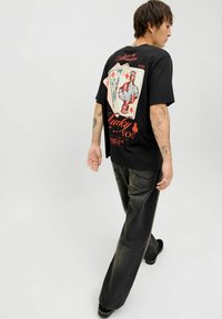 Zwarte katoenen T-shirt met kleurrijk kaartontwerp op de achterkant, voorzien van grafische tekst en patronen; losse pasvorm. Draagt met donkergrijze spijkerbroek.