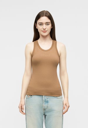 Joven con cabello castaño lacio, vestida con una camiseta marrón sin mangas ajustada y jeans azules claros desgastados, de pie frente a un fondo liso.