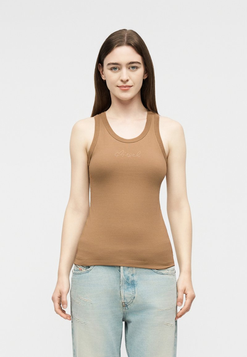 Νεαρή γυναίκα με ίσια καστανά μαλλιά, ντυμένη με εφαρμοστό καφέ μπλουζάκι χωρίς μανίκια και ανοιχτόχρωμα φθαρμένα τζιν, στέκεται μπροστά σε απλό φόντο.