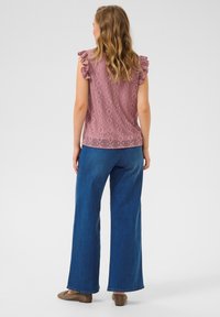 Rozā rakstaina blūze ar ruffles īsām piedurknēm, apvienota ar platām zilo džinsu biksēm un brūniem slip-on apaviem. Mīksts audums ar ģeometrisku dizainu.