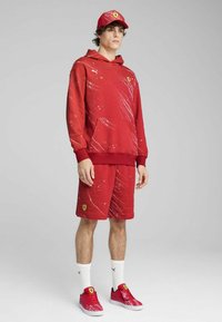 Sweatshirt rouge et shorts avec des motifs éclaboussés blancs, arborant un logo Ferrari. À assortir avec des chaussures rouges et des chaussettes blanches, sur un fond neutre.
