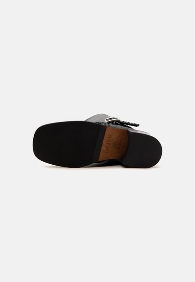 Semelle d'une chaussure en cuir noir avec une bride tressée et un détail de boucle, montrant le nom de la marque et la taille sur une partie en cuir marron.
