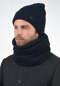 Temno modra pletena kapa z ostrim vrhom in ujemajočim rebrastim vratnim pokrivalom. Ima teksturirano tkanino in diskretno oznako z logotipom.