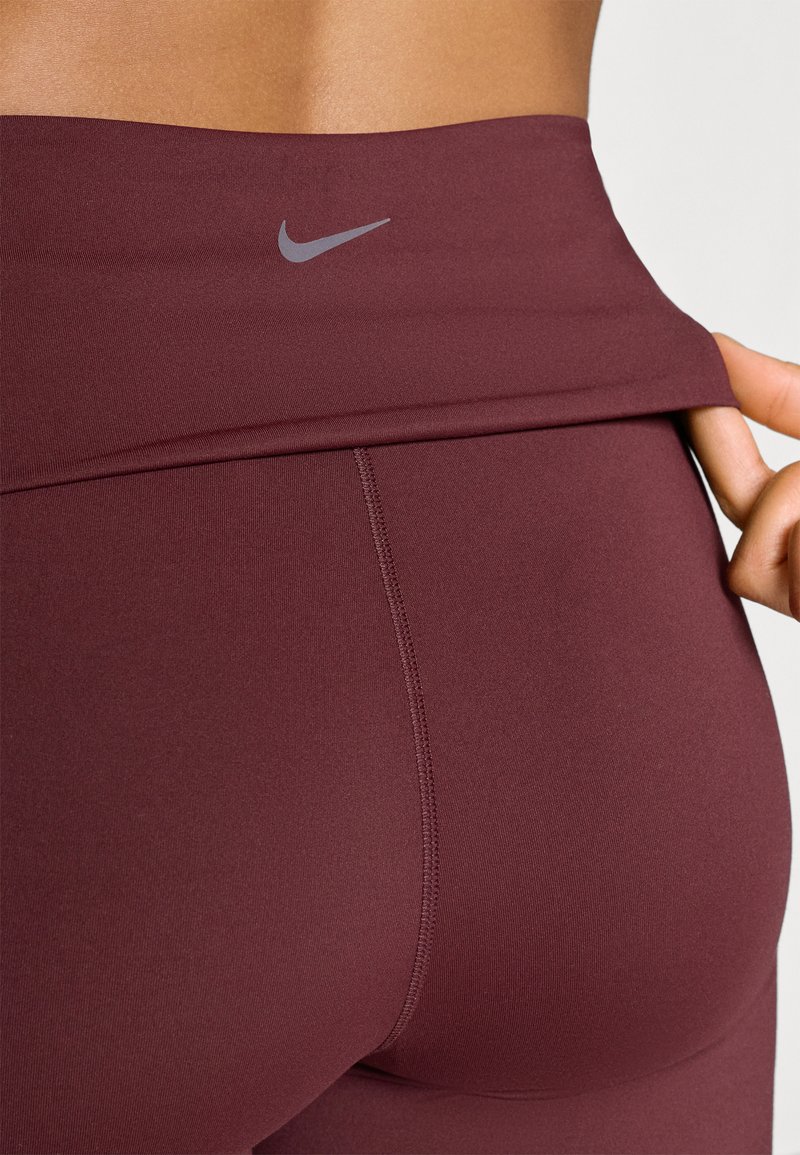 Leggings taille haute bordeaux en tissu extensible. Présente un logo Nike gris sur la ceinture et un design lisse et sans coutures.