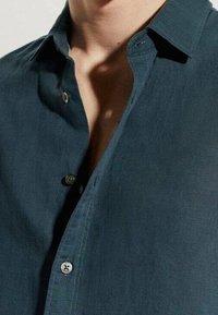 Tummanvihreä paikkapaita, jossa on kaulus, kevyesti kiiltävä pinta ja pehmeä kangas. Näkyvät napit ja avoin etumplikka lisäävät yksityiskohtia.