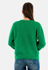 Maglione verde e peloso con scollatura rotonda e spalle scese, indossato sopra jeans blu con cuciture delle tasche distintive.
