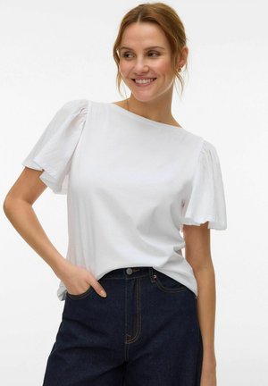 Femme souriante aux cheveux clairs porte un haut blanc à manches bouffantes et un jean bleu foncé, debout avec une main dans la poche devant un fond uni.